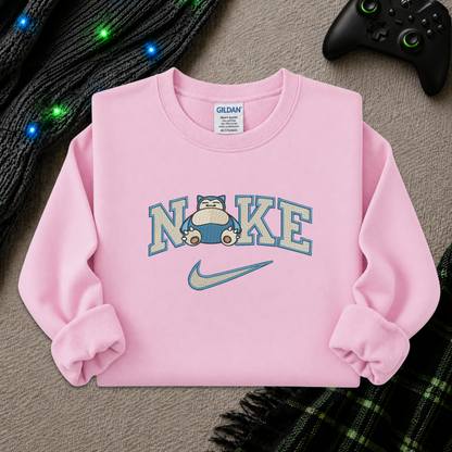 A light pink crewneck sweatshirt featuring a 'NI KE' graphic with a cartoon pokemon snorlax character and a Nike swoosh, styled flat-lay on a textured beige surface with a game controller, knitted sweater, and plaid blanket.
