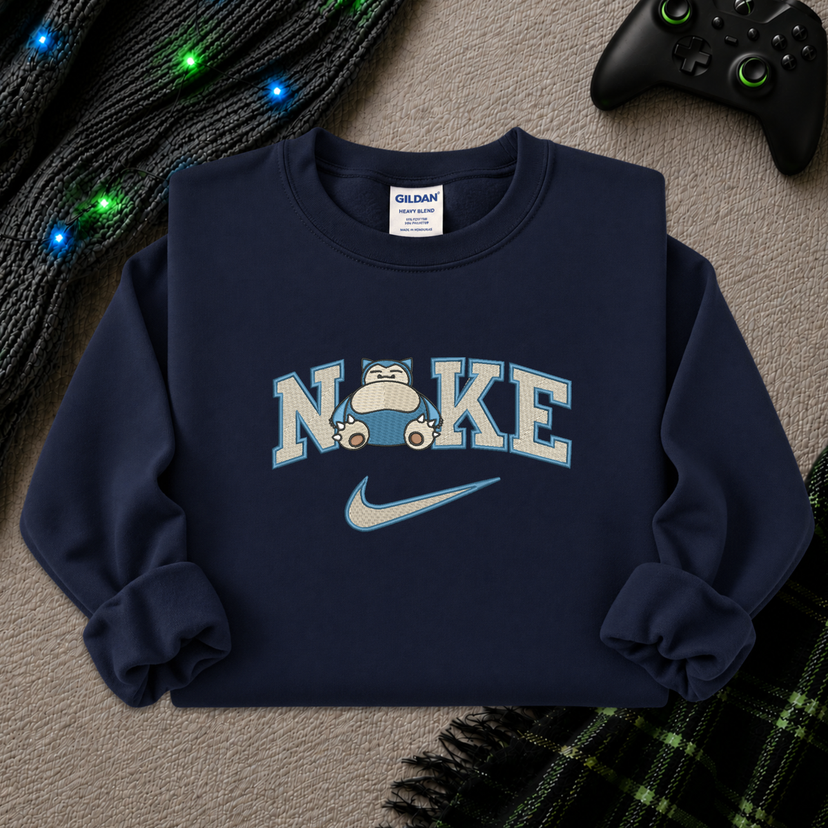 A navy blue crewneck sweatshirt featuring a 'NI KE' graphic with a cartoon pokemon snorlax character and a Nike swoosh, styled flat-lay on a textured beige surface with a game controller, knitted sweater, and plaid blanket.