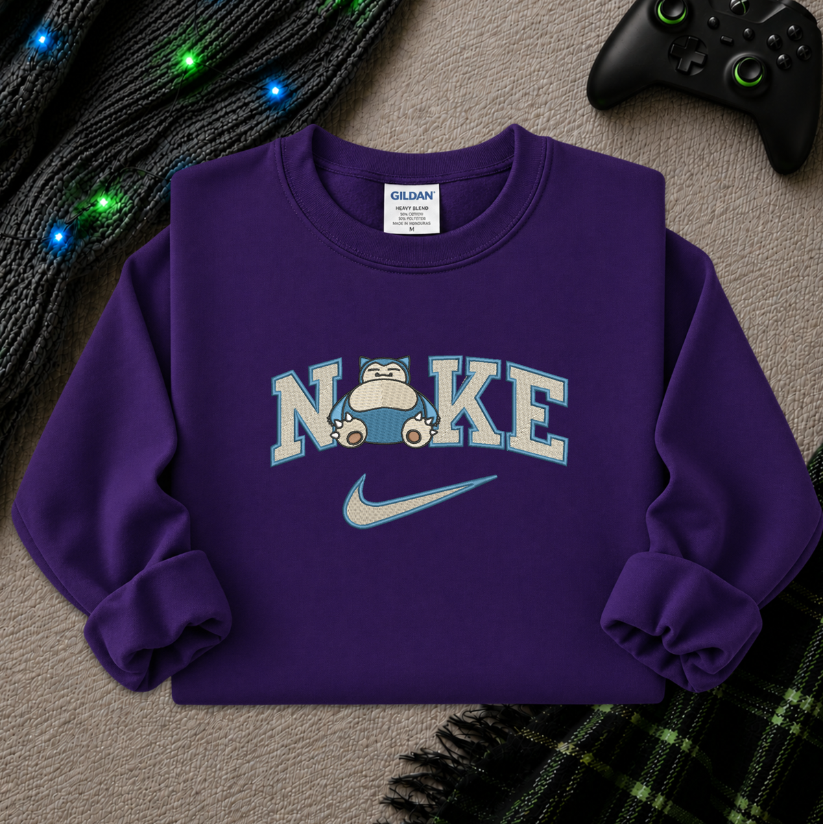 A purple crewneck sweatshirt featuring a 'NI KE' graphic with a cartoon pokemon snorlax character and a Nike swoosh, styled flat-lay on a textured beige surface with a game controller, knitted sweater, and plaid blanket.