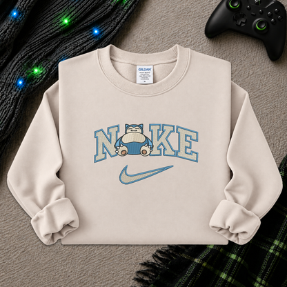 A beige crewneck sweatshirt featuring a 'NI KE' graphic with a cartoon pokemon snorlax character and a Nike swoosh, styled flat-lay on a textured beige surface with a game controller, knitted sweater, and plaid blanket.