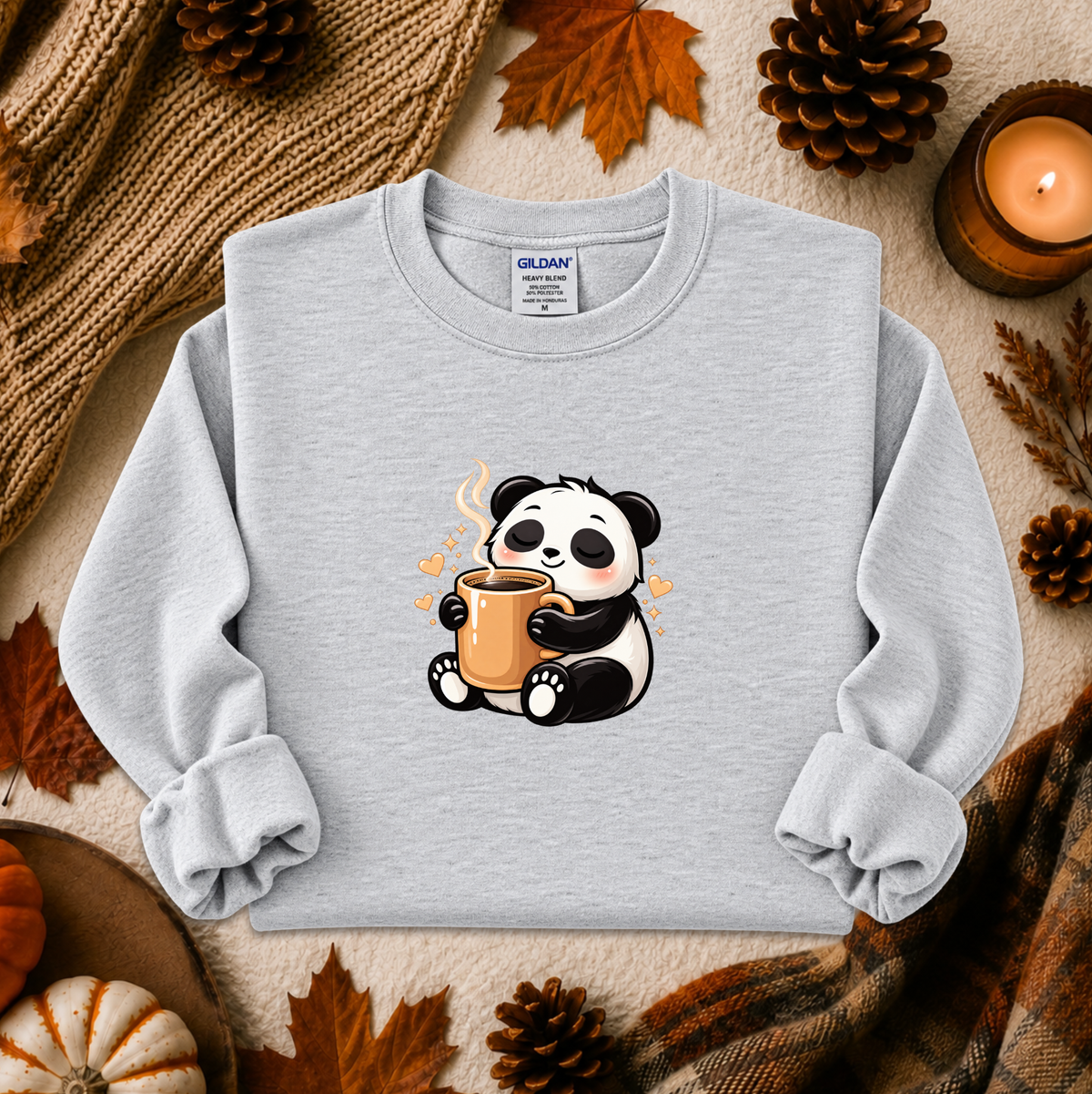 A light heather gray crewneck sweatshirt with a cute cartoon panda holding an orange mug, styled in an autumn flat-lay with pinecones, a candle, and a knit blanket.