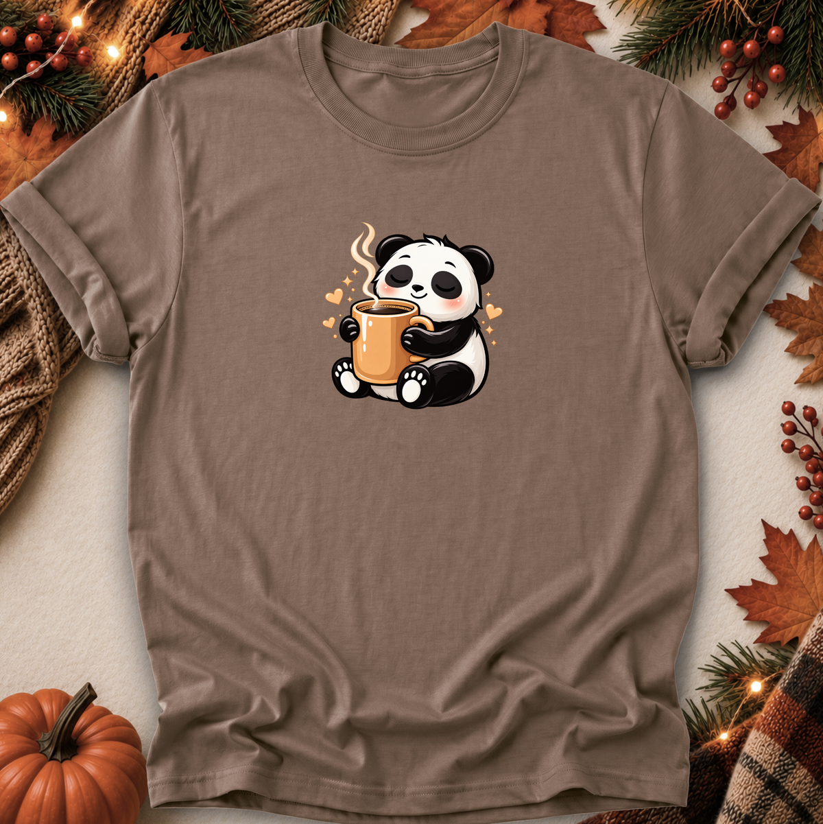 A taupe brown short-sleeve t-shirt with a cute cartoon panda holding a mug, laid flat on a neutral background with autumn-themed props like a pumpkin, pine branches, and leaves.