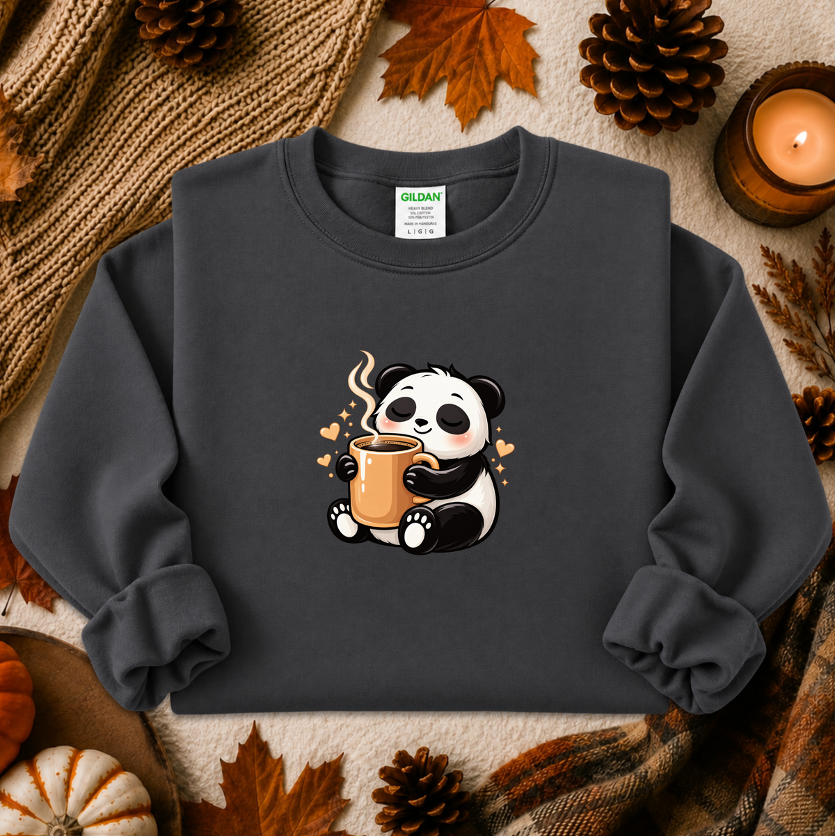 A dark charcoal gray crewneck sweatshirt featuring a cute cartoon panda holding a mug, styled in a cozy autumn flat-lay with pinecones, a knit blanket, and a lit candle. The brand Gildan is visible on the inner neck label.