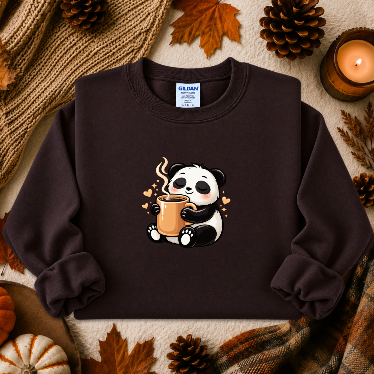 A dark brown crewneck sweatshirt featuring a cute cartoon panda holding a mug, styled in a cozy autumn flat-lay with pinecones, leaves, a candle, and a knit blanket. The brand Gildan is visible on the inner neck label.