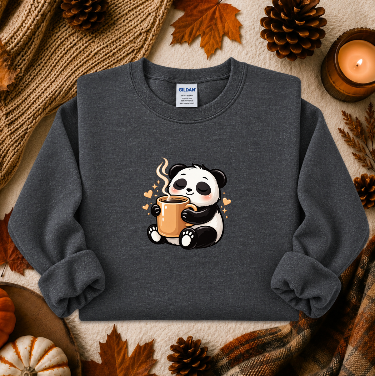 A dark gray crewneck sweatshirt featuring a cute cartoon panda holding a steaming mug, styled in a cozy autumn flat-lay with pinecones, a candle, and a knit blanket.