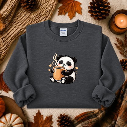 A dark gray crewneck sweatshirt featuring a cute cartoon panda holding a steaming mug, styled in a cozy autumn flat-lay with pinecones, a candle, and a knit blanket.