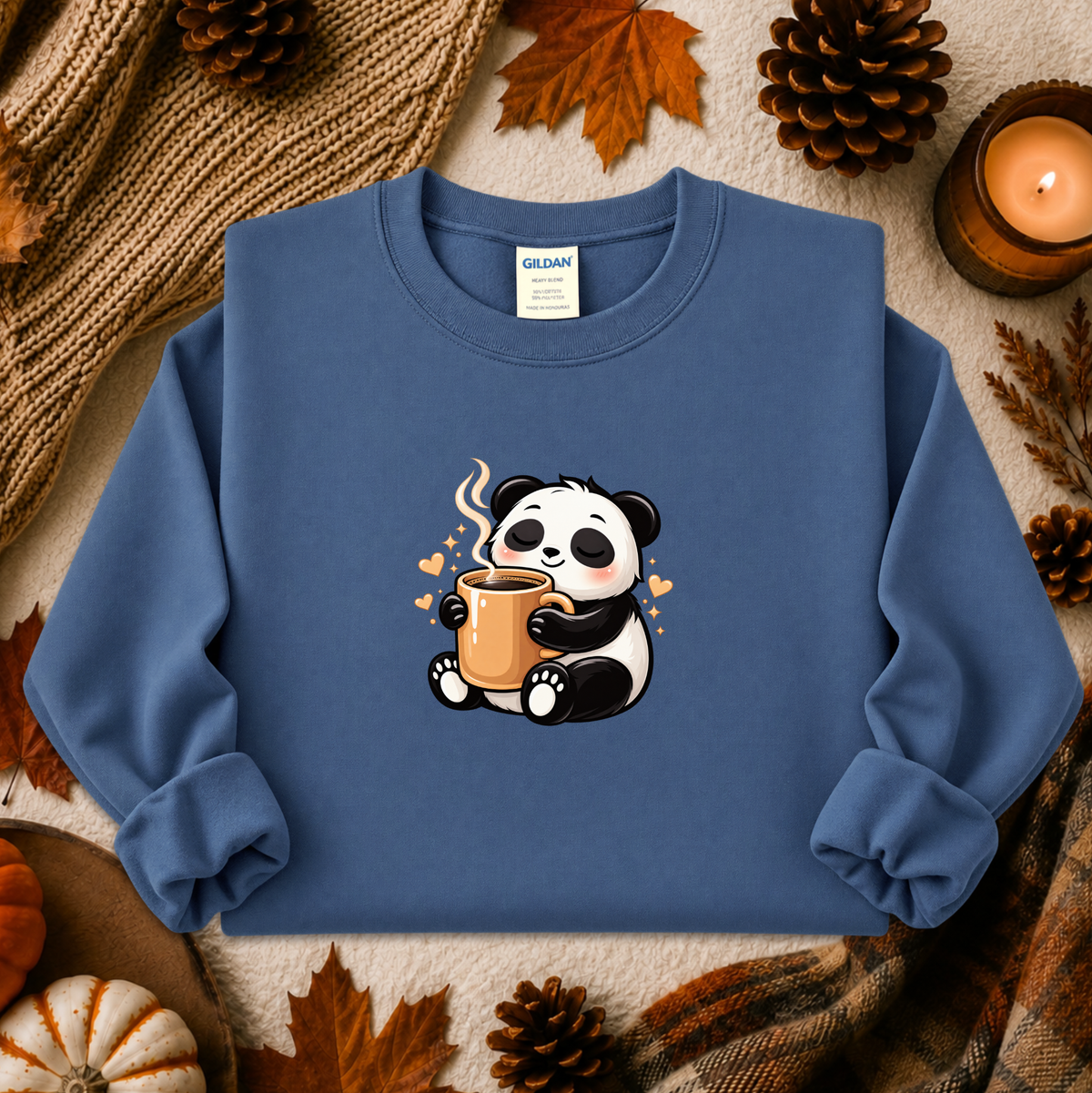 A folded blue crewneck sweatshirt with a cute cartoon panda graphic holding a mug, styled in an autumn flat-lay with pinecones, leaves, a candle, and a knit blanket. The neck label shows the brand Gildan.