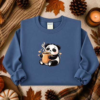 A folded blue crewneck sweatshirt with a cute cartoon panda graphic holding a mug, styled in an autumn flat-lay with pinecones, leaves, a candle, and a knit blanket. The neck label shows the brand Gildan.