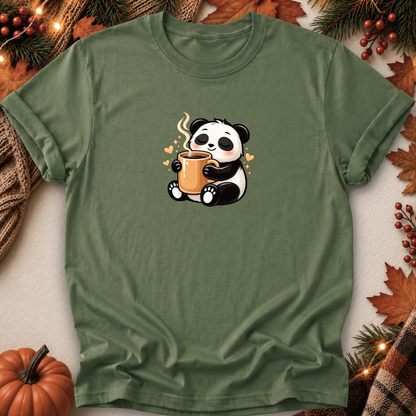 An olive green short-sleeve t-shirt with a cute cartoon panda holding an orange mug, styled in a flat-lay with autumn decorations like a pumpkin, pine branches, and leaves.