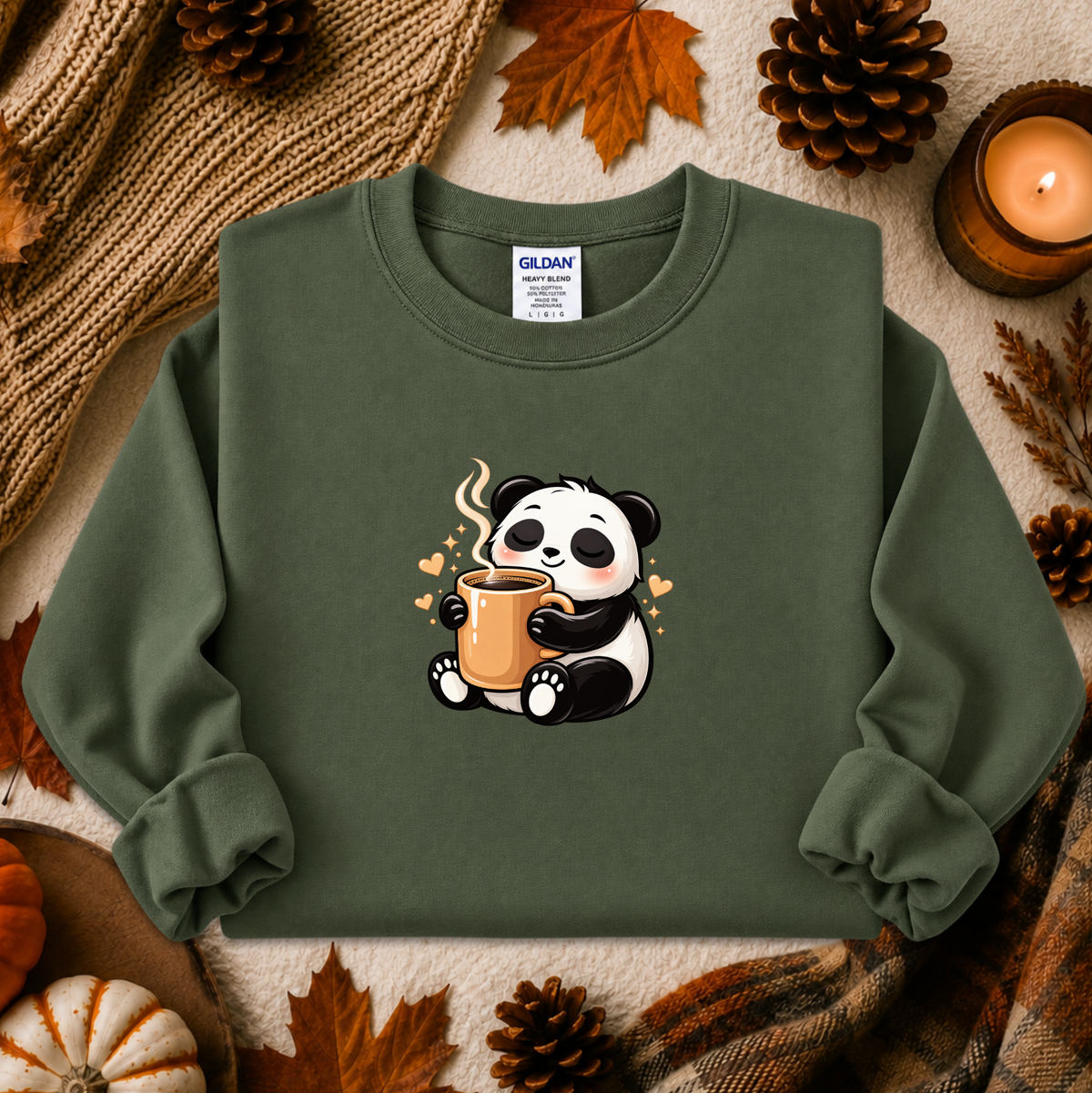An olive green crewneck sweatshirt by Gildan, featuring a cute cartoon panda holding a mug, styled in a cozy autumn flat-lay with pinecones, a candle, and a knit blanket.