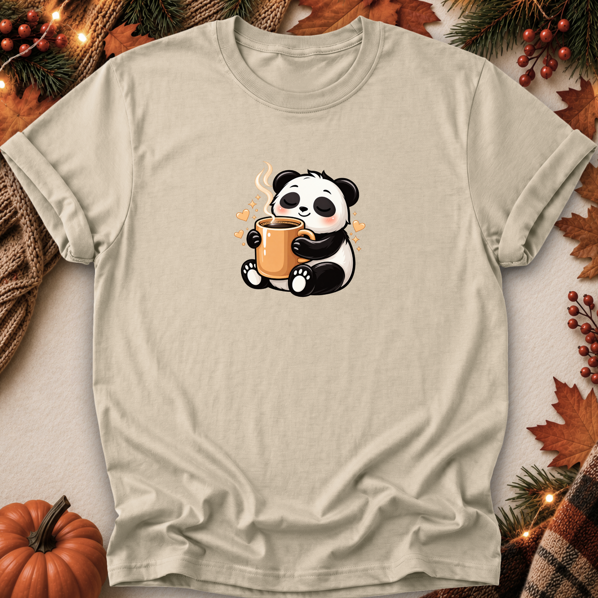 A light beige crewneck t-shirt featuring a cute cartoon panda holding an orange mug, styled in a flat-lay with autumn decorations including a small pumpkin, pine branches, and leaves.