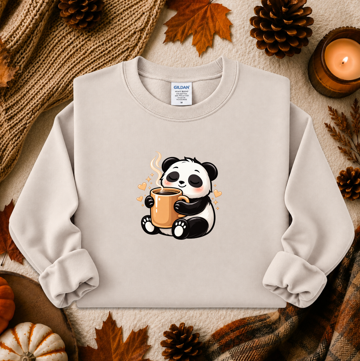 A light beige crewneck sweatshirt featuring a cute cartoon panda holding an orange mug, styled in a cozy flat-lay with autumn leaves, pinecones, a candle, and a knitted scarf.