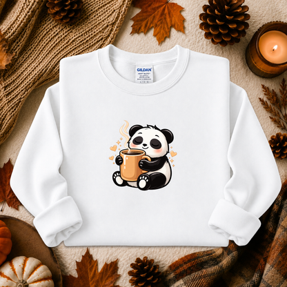 A white long-sleeve crewneck sweatshirt by Gildan, featuring a cute cartoon panda graphic holding an orange mug. The sweatshirt is displayed in a flat-lay arrangement with autumn-themed props including pinecones, a lit candle, and a knitted scarf.