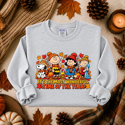 A light gray long-sleeve crewneck sweatshirt featuring a colorful cartoon graphic of Peanuts characters in autumn clothing with the text 'IT'S THE MOST WONDERFUL TIME OF THE YEAR'. The sweatshirt is styled in a flat lay with fall-themed props including pinecones, leaves, a candle, and a knit blanket.