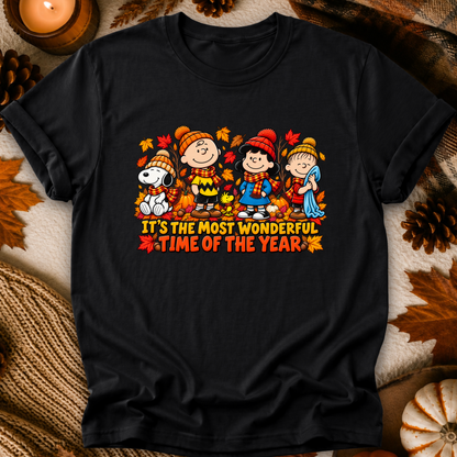 A black short-sleeve graphic t-shirt featuring the Peanuts characters Snoopy, Charlie Brown, Woodstock, and Lucy in autumn clothing with the text 'IT'S THE MOST WONDERFUL TIME OF THE YEAR'. The shirt is displayed in a flat-lay setting with autumn props like pinecones, leaves, and a candle.