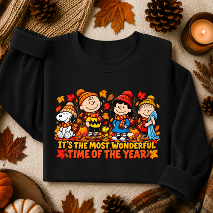 A black long-sleeve sweatshirt laid flat on a cozy autumn-themed background with pinecones, leaves, a candle, and plaid fabric. The front has a colorful cartoon graphic of children and a dog among fall leaves, with the text 'IT'S THE MOST WONDERFUL TIME OF THE YEAR' below.