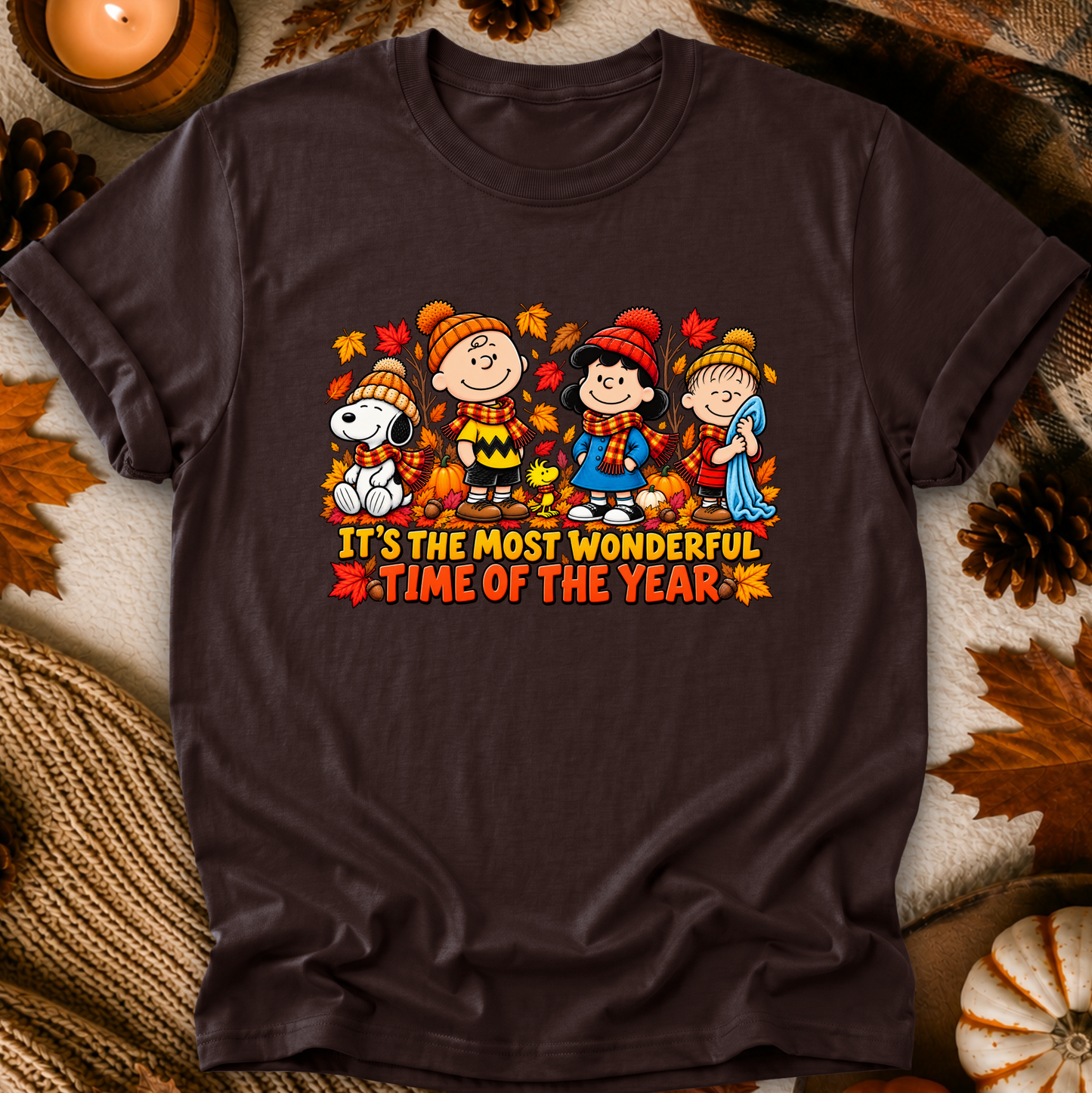 A dark brown short-sleeve t-shirt featuring a colorful autumn-themed graphic of Peanuts characters with the text 'IT'S THE MOST WONDERFUL TIME OF THE YEAR'. The shirt is displayed in a flat-lay setting surrounded by fall props including pine cones, a candle, and a knitted blanket.
