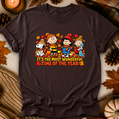 A dark brown short-sleeve t-shirt featuring a colorful autumn-themed graphic of Peanuts characters with the text 'IT'S THE MOST WONDERFUL TIME OF THE YEAR'. The shirt is displayed in a flat-lay setting surrounded by fall props including pine cones, a candle, and a knitted blanket.