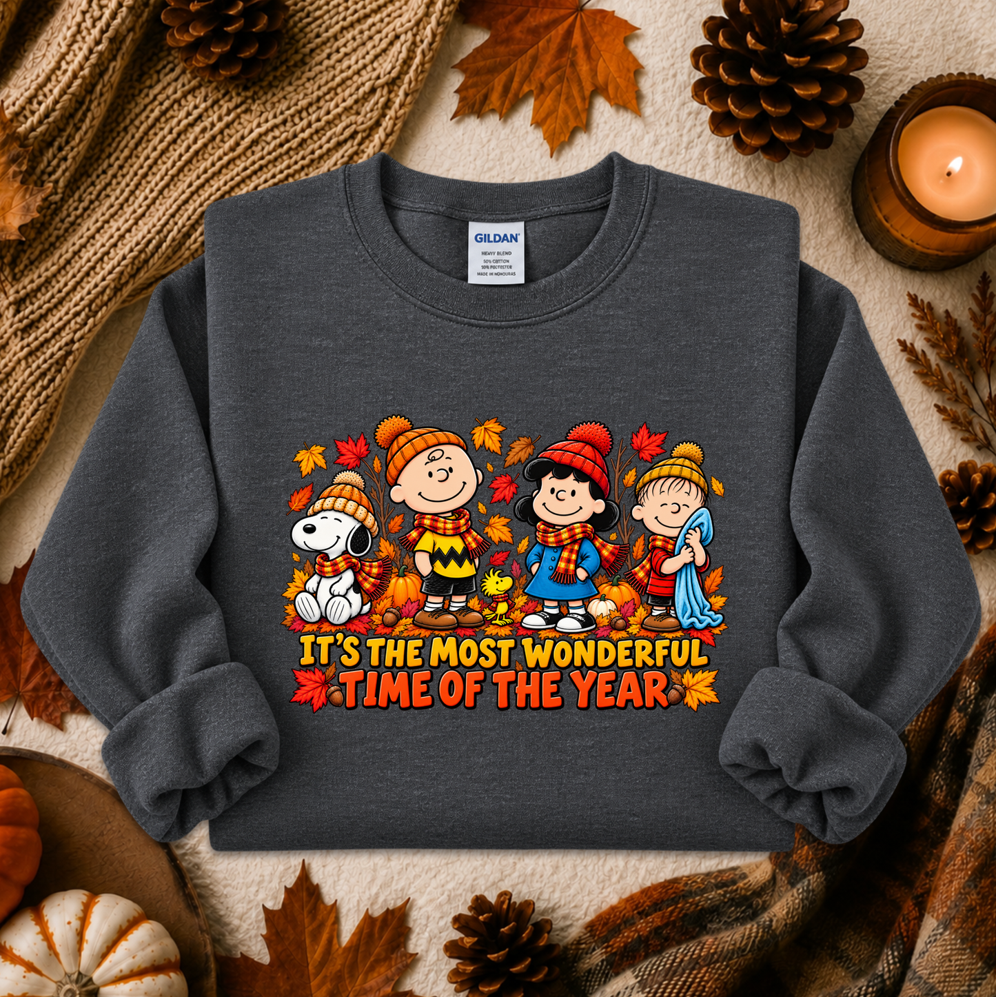 A dark gray crewneck sweatshirt featuring a colorful graphic of the Peanuts characters in autumn clothing with the text 'IT'S THE MOST WONDERFUL TIME OF THE YEAR'. The sweatshirt is styled in a flat-lay arrangement with fall-themed props including pinecones, a candle, a knit blanket, and a small pumpkin.