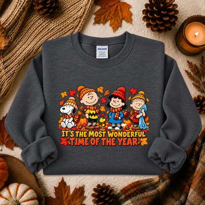 A dark gray crewneck sweatshirt featuring a colorful graphic of the Peanuts characters in autumn clothing with the text 'IT'S THE MOST WONDERFUL TIME OF THE YEAR'. The sweatshirt is styled in a flat-lay arrangement with fall-themed props including pinecones, a candle, a knit blanket, and a small pumpkin.