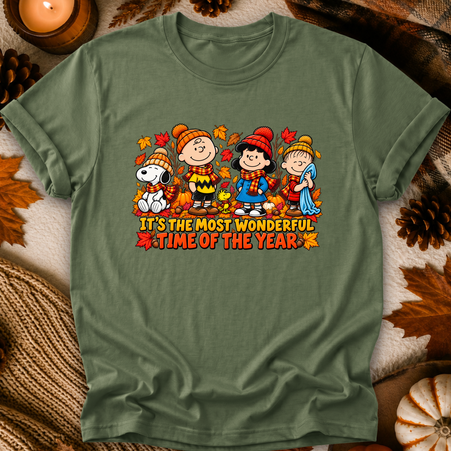 An olive green crewneck t-shirt featuring a colorful graphic of Peanuts characters (Snoopy, Charlie Brown, Lucy, and Sally) in autumn clothing with the text 'IT'S THE MOST WONDERFUL TIME OF THE YEAR'. The shirt is displayed in a flat-lay setting with fall-themed props like pinecones, a candle, and a knit blanket.