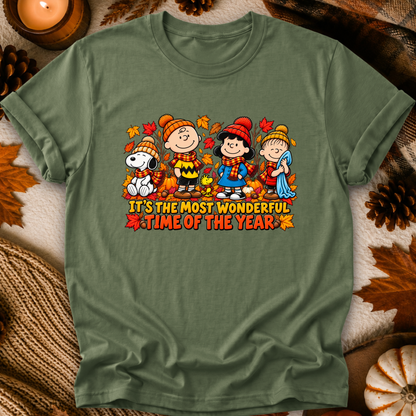 An olive green crewneck t-shirt featuring a colorful graphic of Peanuts characters (Snoopy, Charlie Brown, Lucy, and Sally) in autumn clothing with the text 'IT'S THE MOST WONDERFUL TIME OF THE YEAR'. The shirt is displayed in a flat-lay setting with fall-themed props like pinecones, a candle, and a knit blanket.