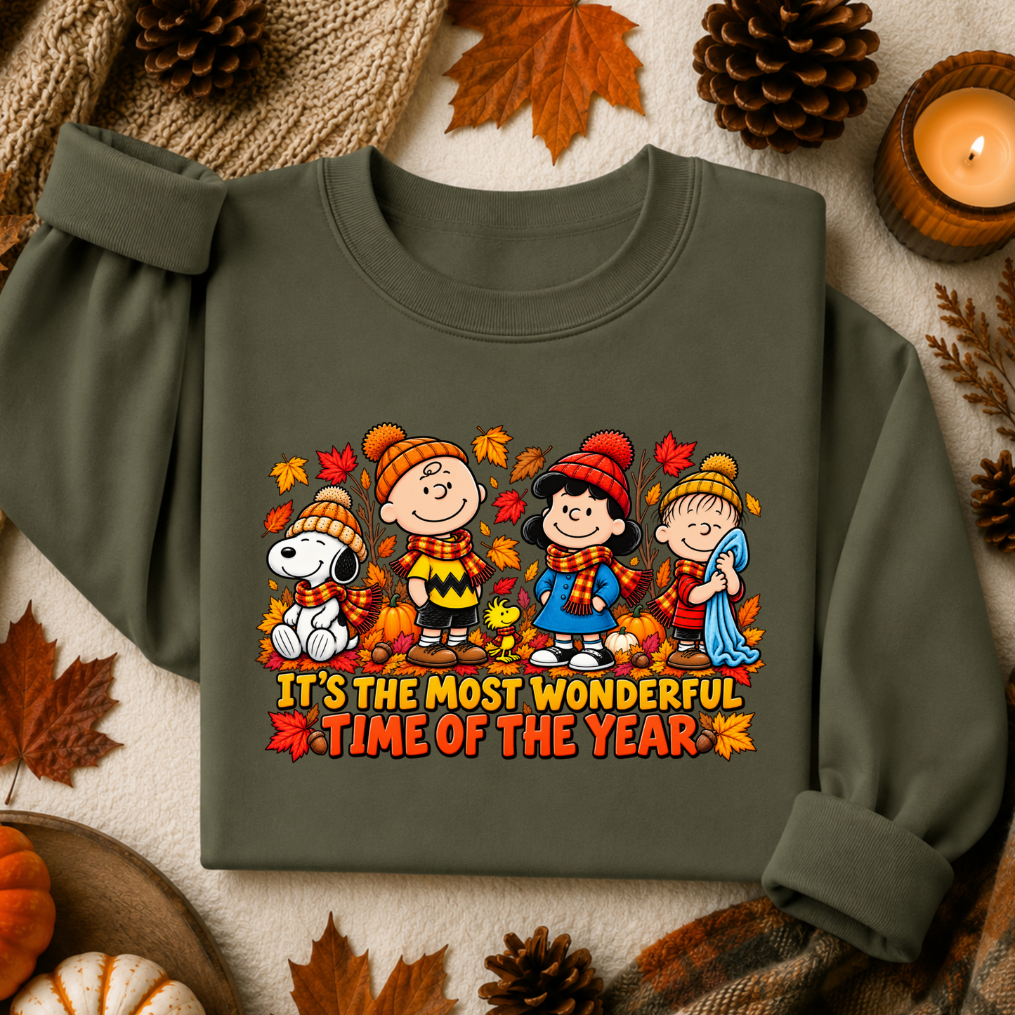 An olive green long-sleeve sweatshirt featuring a colorful graphic of Peanuts characters in autumn clothing with the text 'IT'S THE MOST WONDERFUL TIME OF THE YEAR'. The sweatshirt is styled in a flat lay with fall-themed props including pinecones, autumn leaves, a candle, and a small pumpkin on a textured cream fabric background.