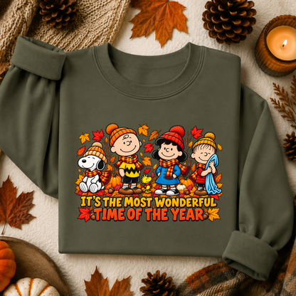 An olive green long-sleeve sweatshirt featuring a colorful graphic of Peanuts characters in autumn clothing with the text 'IT'S THE MOST WONDERFUL TIME OF THE YEAR'. The sweatshirt is styled in a flat lay with fall-themed props including pinecones, autumn leaves, a candle, and a small pumpkin on a textured cream fabric background.