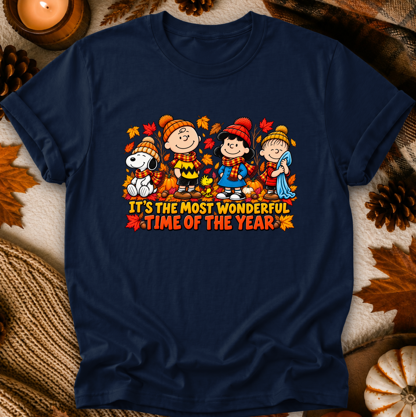 A navy blue graphic t-shirt featuring a Peanuts autumn-themed illustration with the text 'IT'S THE MOST WONDERFUL TIME OF THE YEAR'. The shirt is displayed in a flat-lay setting surrounded by fall-themed props including pinecones, a lit candle, and a knitted blanket.