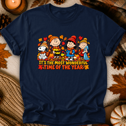 A navy blue graphic t-shirt featuring a Peanuts autumn-themed illustration with the text 'IT'S THE MOST WONDERFUL TIME OF THE YEAR'. The shirt is displayed in a flat-lay setting surrounded by fall-themed props including pinecones, a lit candle, and a knitted blanket.