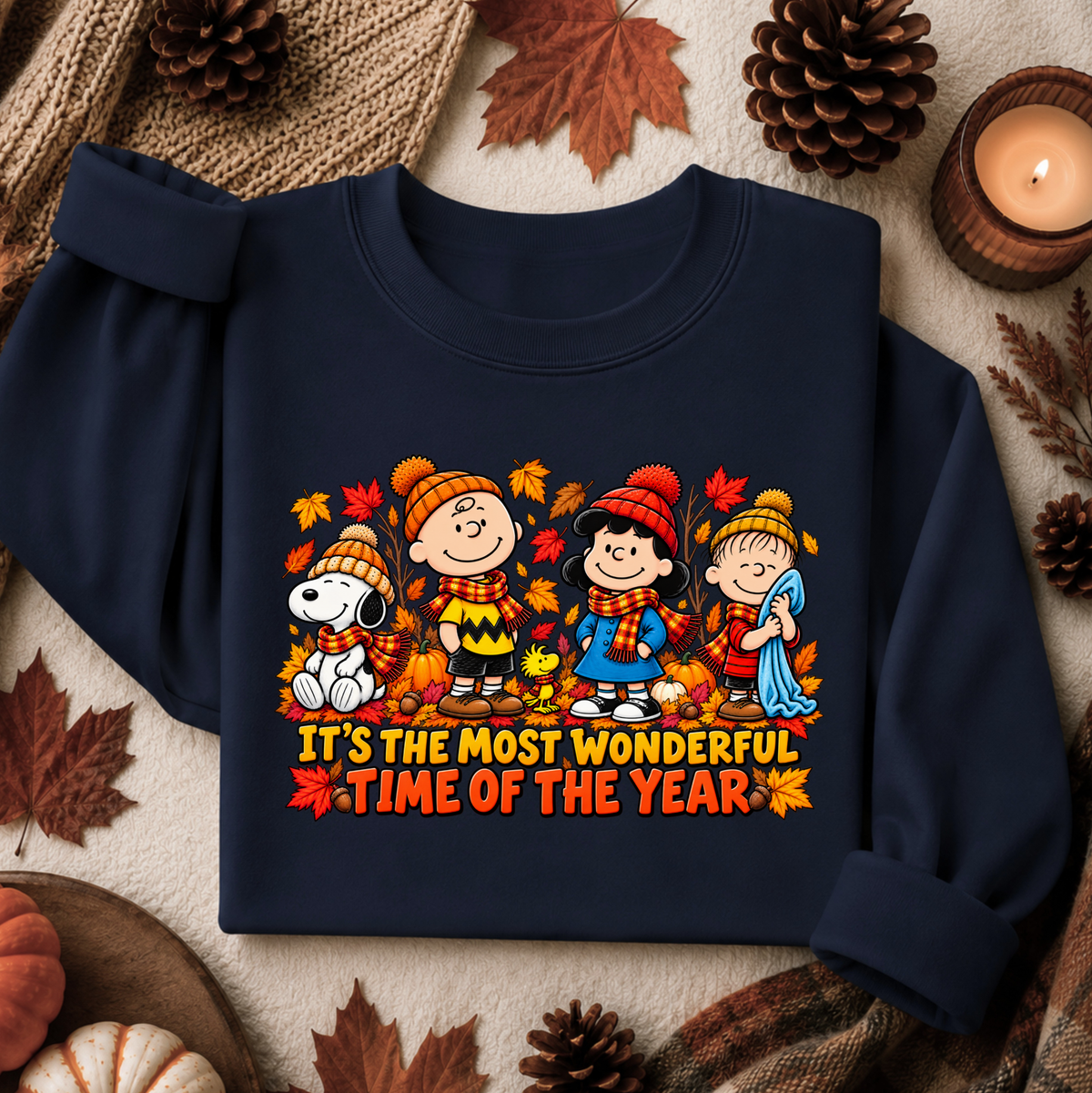 A navy blue sweatshirt laid flat on a cozy autumn-themed background with pinecones, leaves, and a lit candle. The sweatshirt features a colorful graphic of Peanuts characters wearing fall clothing, surrounded by leaves, with the text 'IT'S THE MOST WONDERFUL TIME OF THE YEAR' printed below.