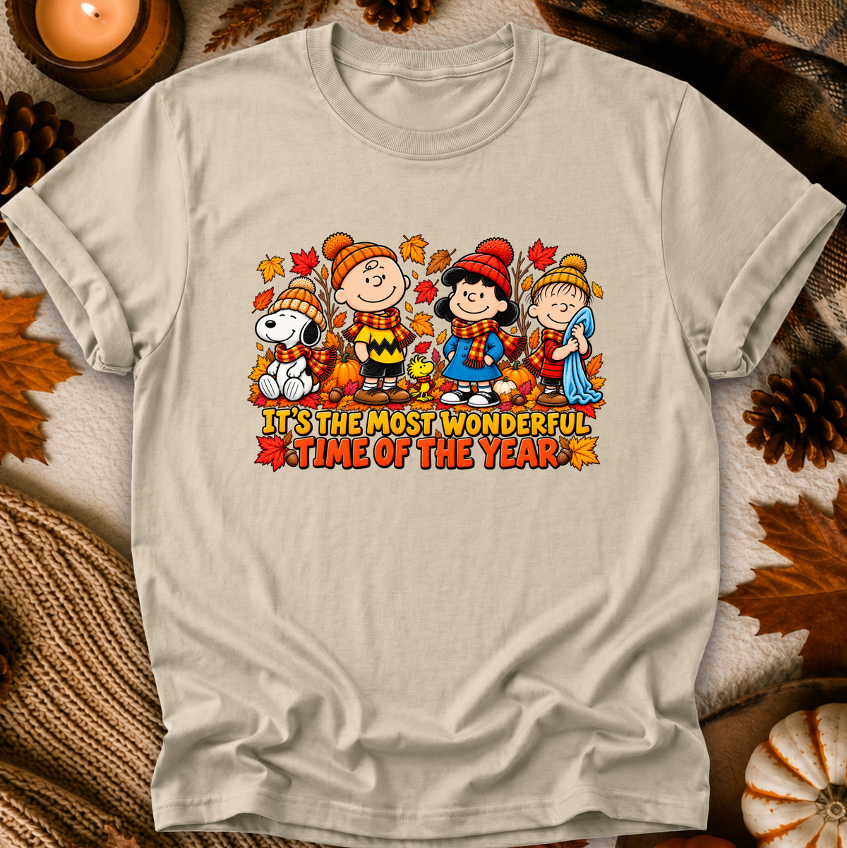 A light beige graphic t-shirt featuring Peanuts characters in autumn clothing with the text 'IT'S THE MOST WONDERFUL TIME OF THE YEAR'. The shirt is displayed in a flat-lay setting with fall-themed props like pinecones, a candle, and a knit blanket.