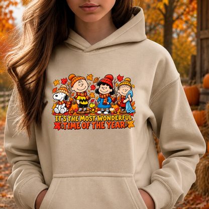 A woman wearing a beige hoodie with a colorful graphic of Peanuts characters and the text 'IT'S THE MOST WONDERFUL TIME OF THE YEAR', photographed outdoors in an autumn setting with pumpkins and fall foliage.