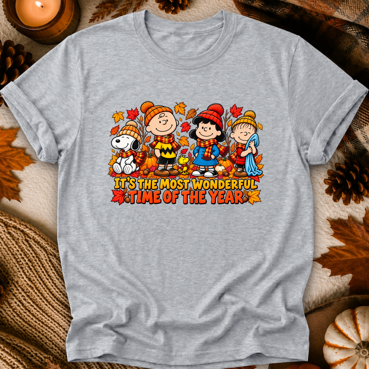 A light heather gray crewneck t-shirt featuring a colorful graphic of Peanuts characters in autumn outfits with the text 'IT'S THE MOST WONDERFUL TIME OF THE YEAR'. The shirt is displayed in a flat-lay setting with fall-themed props like pinecones, a candle, and a pumpkin.
