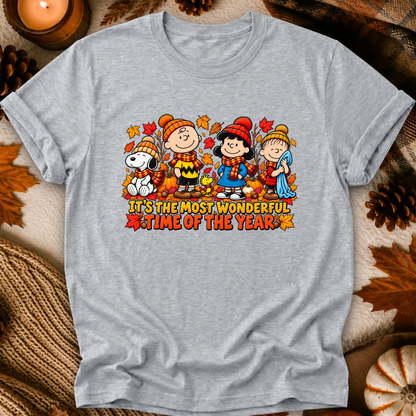 A light heather gray crewneck t-shirt featuring a colorful graphic of Peanuts characters in autumn outfits with the text 'IT'S THE MOST WONDERFUL TIME OF THE YEAR'. The shirt is displayed in a flat-lay setting with fall-themed props like pinecones, a candle, and a pumpkin.