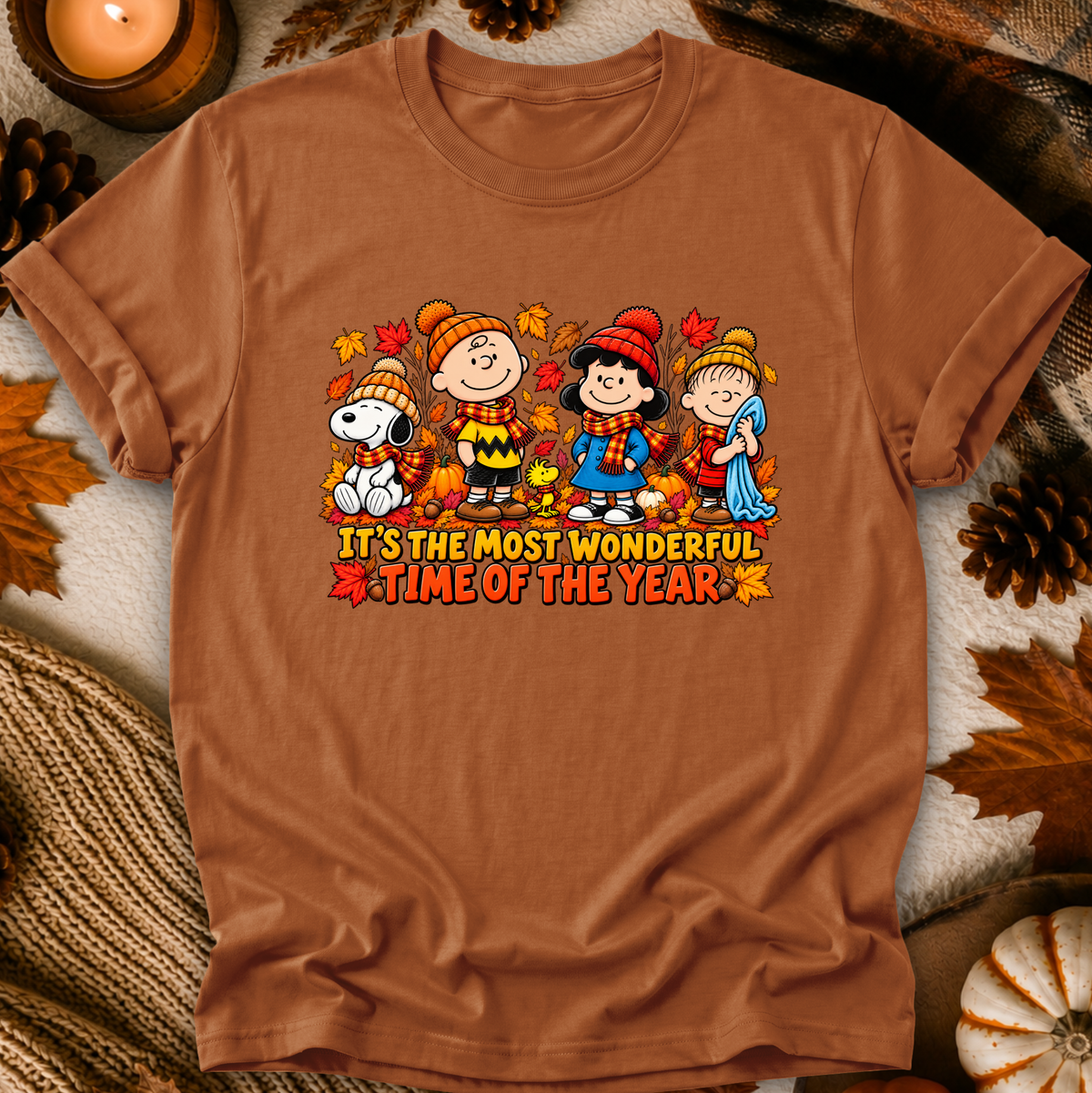 A brown short-sleeve graphic T-shirt laid flat on a cozy autumn-themed background with pinecones, leaves, a candle, and knit fabric. The shirt features a colorful cartoon print of Peanuts characters wearing fall clothing with the text 'IT'S THE MOST WONDERFUL TIME OF THE YEAR'.