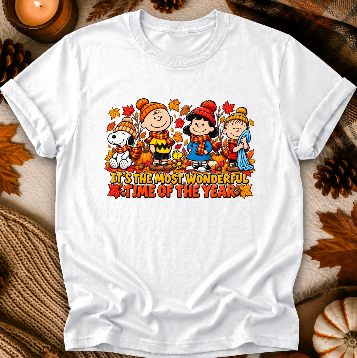 A white short-sleeve crewneck t-shirt featuring a colorful graphic of Peanuts characters in autumn outfits with the text 'IT'S THE MOST WONDERFUL TIME OF THE YEAR'. The shirt is displayed in a flat-lay setting with cozy fall props like pinecones, a candle, and a knit blanket.