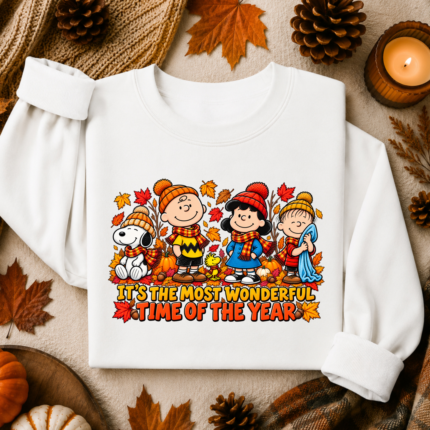 A white long-sleeve sweatshirt laid flat on a cozy autumn-themed background with pinecones, leaves, and a candle. The front has a colorful cartoon graphic of children in fall clothing with the text 'IT'S THE MOST WONDERFUL TIME OF THE YEAR'.