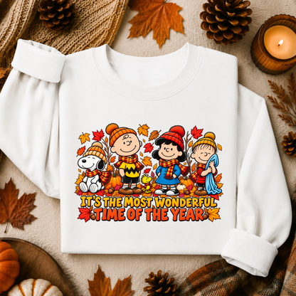 A white long-sleeve sweatshirt laid flat on a cozy autumn-themed background with pinecones, leaves, and a candle. The front has a colorful cartoon graphic of children in fall clothing with the text 'IT'S THE MOST WONDERFUL TIME OF THE YEAR'.