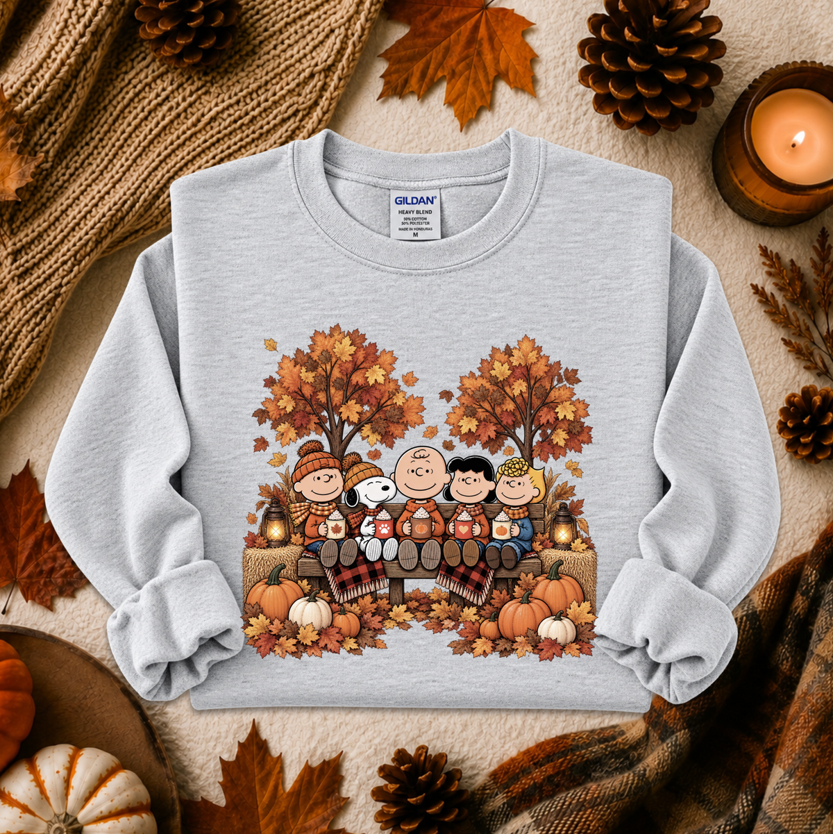 A light gray crewneck sweatshirt featuring a colorful cartoon graphic of Snoopy and his friends sitting in a pumpkin patch surrounded by autumn trees and pumpkins. The sweatshirt is styled in a cozy flat-lay arrangement with a knitted scarf, pinecones, fall leaves, and a lit candle.