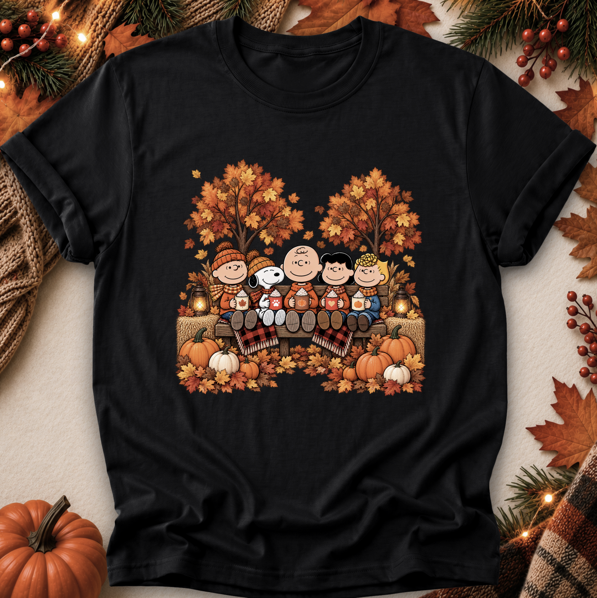 A black short-sleeve graphic T-shirt laid flat on a neutral surface, surrounded by autumn-themed props including pumpkins, leaves, pine branches, and string lights. The shirt features a colorful printed illustration of the Peanuts characters sitting by a fire in the fall.