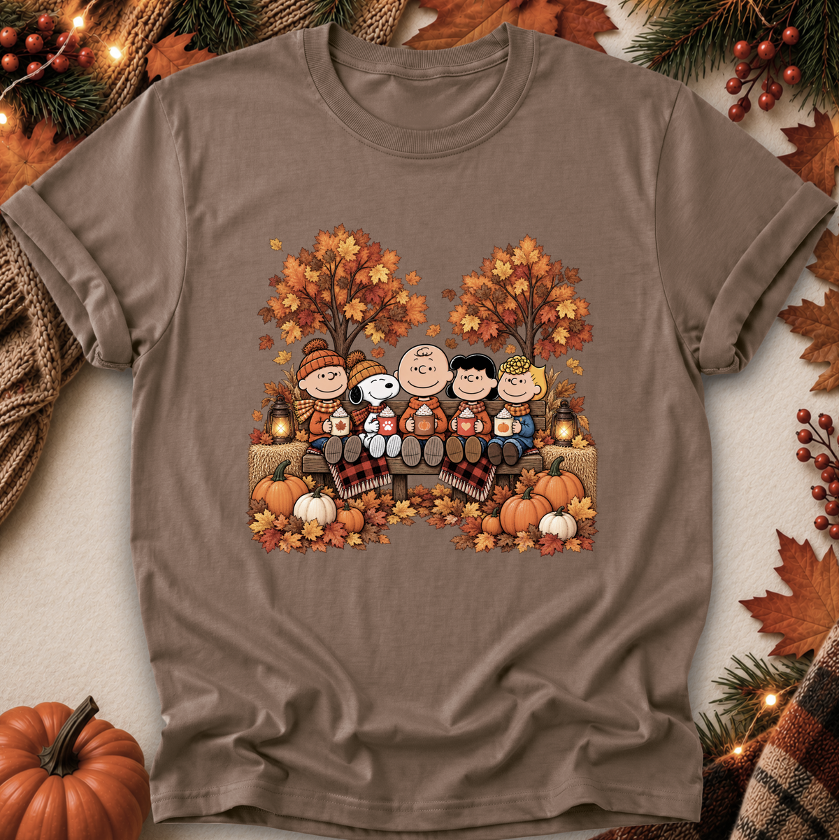 A taupe brown short-sleeve t-shirt featuring a colorful graphic of the Peanuts characters in autumn clothing, surrounded by pumpkins, fall leaves, and pine branches.