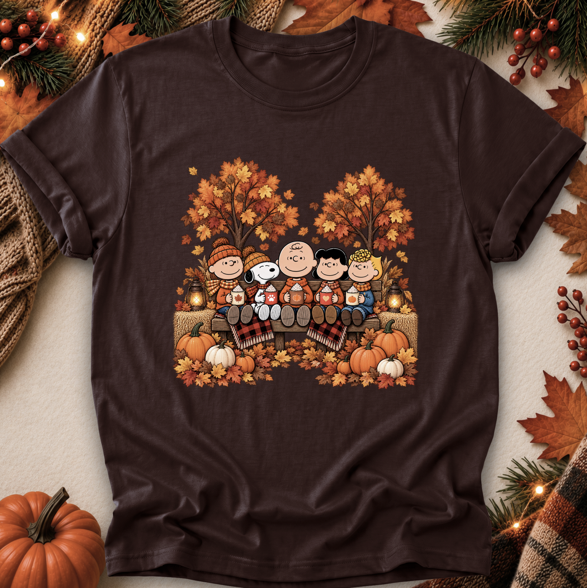 A dark brown short-sleeve crewneck t-shirt featuring a colorful graphic of the Peanuts characters sitting on a bench surrounded by autumn trees, pumpkins, and fall leaves. The shirt is displayed in a flat-lay arrangement with seasonal props including a small pumpkin, pine branches, and leaves.