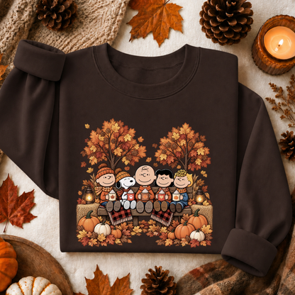 A dark brown crewneck sweatshirt featuring a colorful cartoon graphic of Peanuts characters sitting on a piano surrounded by autumn trees, pumpkins, and leaves. The sweatshirt is styled in a flat lay with seasonal props including pinecones, a candle, and fall leaves on a neutral fabric background.