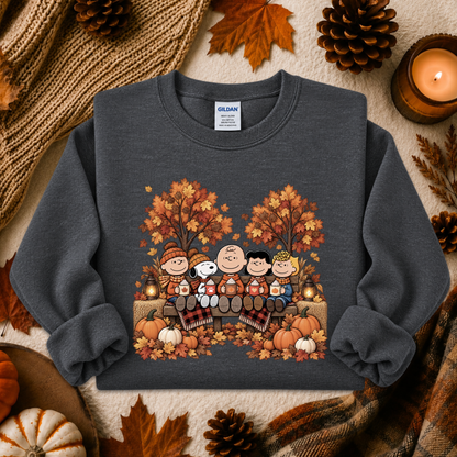 A dark gray crewneck sweatshirt featuring a colorful graphic of the Peanuts holiday family sitting on a bench surrounded by autumn trees, pumpkins, and leaves. The sweatshirt is styled in a cozy flat-lay arrangement with a knitted scarf, pinecones, fall leaves, and a lit candle.