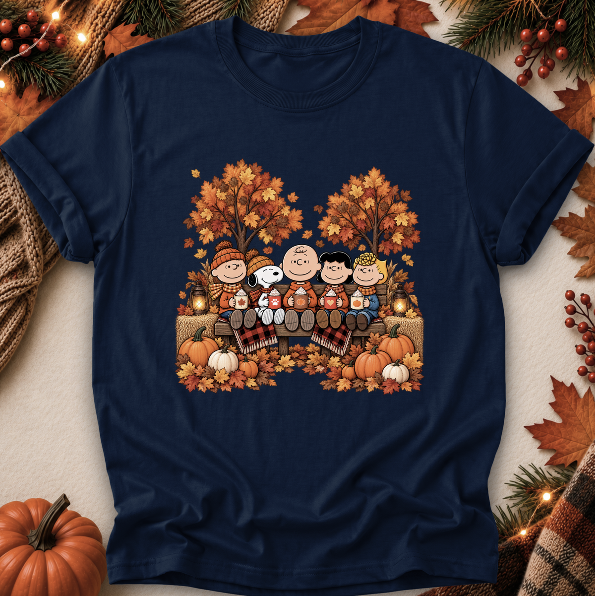 A navy blue crewneck t-shirt featuring a colorful graphic of the Peanuts characters Snoopy, Charlie Brown, and Woodstock sitting on a bench surrounded by autumn trees, pumpkins, and fall leaves. The shirt is displayed in a flat-lay arrangement with seasonal props including pine branches, small pumpkins, and warm string lights.