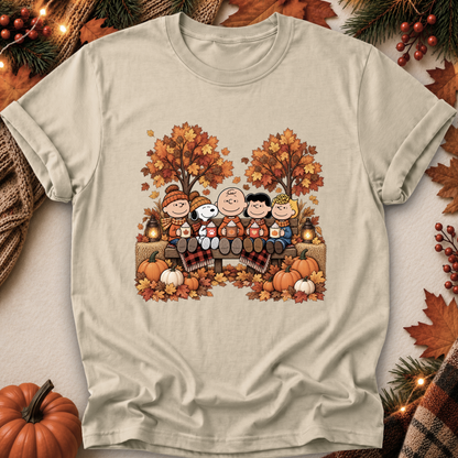 A light beige graphic t-shirt featuring a cartoon fall scene with characters sitting on a bench surrounded by pumpkins, autumn trees, and leaves. The shirt is displayed in a flat-lay arrangement with seasonal props like a pumpkin, pine branches, and a plaid scarf.