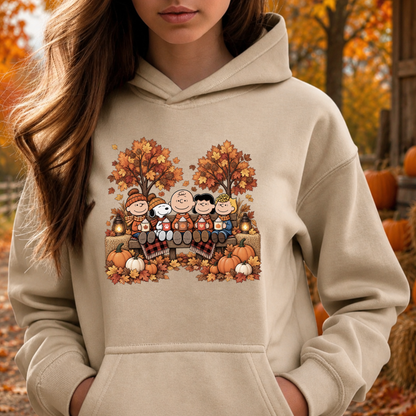 A woman wearing a beige hoodie with a colorful autumn-themed graphic print of Peanuts characters sitting on a bench surrounded by pumpkins and fall leaves. The background shows an outdoor autumn scene with trees and pumpkins.