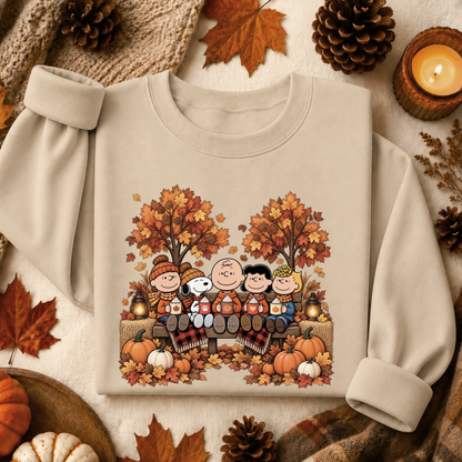 A cream-colored long-sleeve sweatshirt featuring a colorful graphic of the Peanuts characters sitting on a bench surrounded by autumn trees and pumpkins. The sweatshirt is styled in a cozy flat-lay arrangement with fall leaves, pinecones, a candle, and a knit scarf.