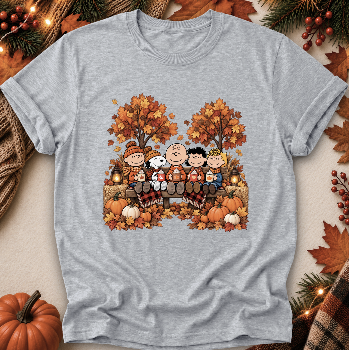 A heather gray short-sleeve t-shirt featuring a colorful autumn-themed graphic of Peanuts characters sitting by a campfire. The shirt is displayed in a flat-lay setting surrounded by seasonal props including pumpkins, fall leaves, pine branches, and string lights.